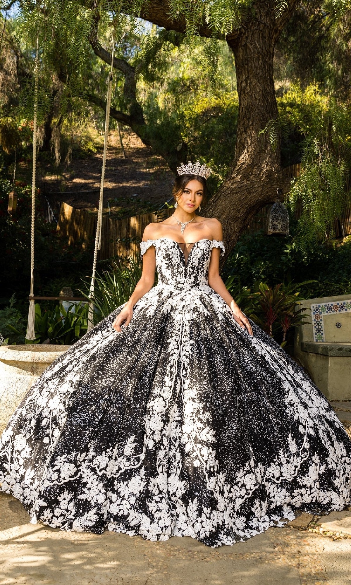 A woman in the Cinderella Couture 8070J ornate black and white off-the-shoulder quinceanera dress with floral lace embroidery and a jeweled crown stands outdoors before a large tree.